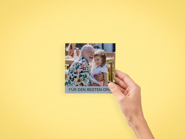 Ein Fotobuch mit den Maßen 13x13 cm wird von einer Hand gehalten. Das Cover zeigt einen Großvater mit seiner Enkelin. 