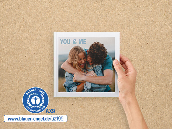 Ein Fotobuch im Format 20x20 wird von einer Hand gehalten. Auf dem Cover ist ein Paar zu sehen. Daneben ist der Blaue Engel.