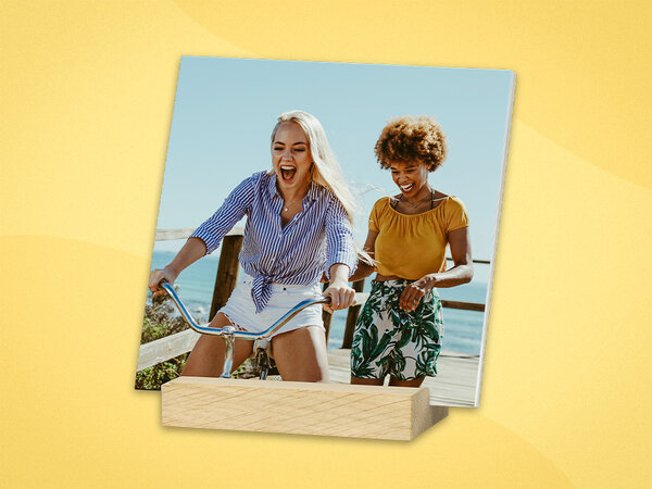 Mehrere quadratische Fotos stehen frei in einem Holzstandfuß. Das vorderste Foto zeigt zwei Frauen, die miteinander Lachen.